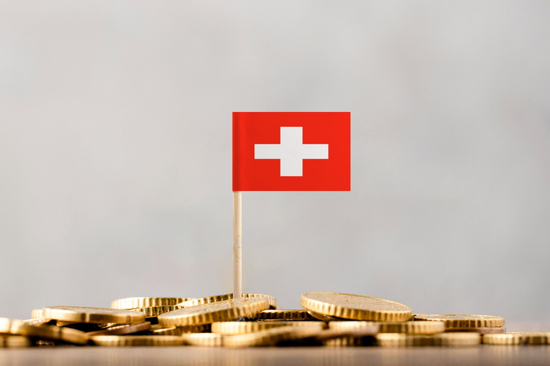 Small miniature Swiss flag planted atop a pile of gold coins, symbolizing wealth management Switzerland, Swiss franc currency diversification, and foreign asset custody for U.S. citizens.